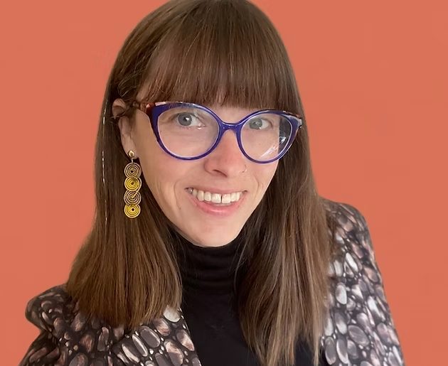 Dr. Laura Colket smiling, with long brown hair, bangs and blue cat-eye glasses, against a coral background
